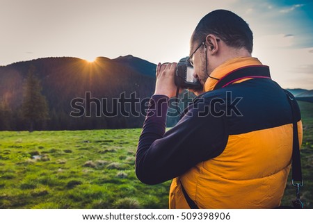 Nature photographer tourist with camera taking a photo in the mountains. Dreamy sunset landscape, spring green meadow and mountain top in the bsckground. Back view