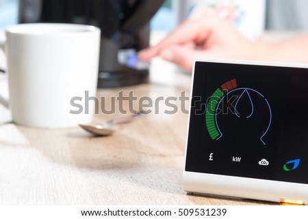 A smart meter is displayed on a  wooden surface near mug and spoon and a kettle which is being switched on by a hand. The meter is giving a digital reading of energy consumption. COPY SPACE Royalty-Free Stock Photo #509531239