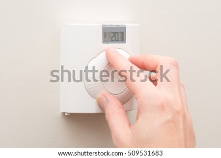 A hand turns the dial on a white wall mounted thermostat reading 20 degrees room temperature Royalty-Free Stock Photo #509531683