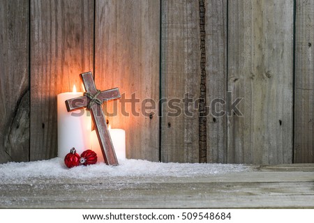White holiday candles glowing behind wooden cross with red Christmas ornaments in snow by antique rustic wood background; Christmas and religious background with copy space