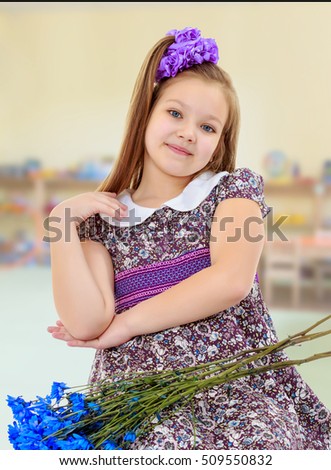 Happy little girl with a big purple bow on her head , and fancy dress. In the lap of the girls lay a bouquet of blue flowers.In the children's room with toys
