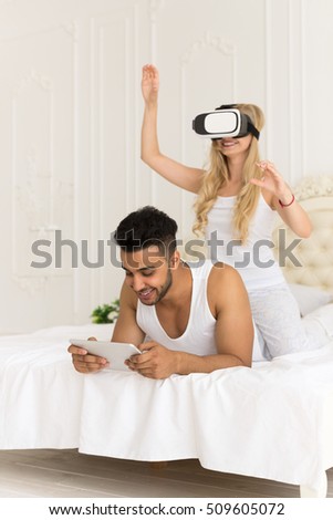 Young Couple In Bed, Hispanic Man Using Cell Smart Phone Woman Wear Virtual Reality Digital Glasses Headset, Lovers In Bedroom