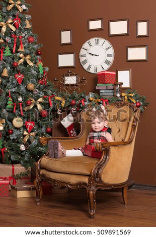 Charming baby girl with a gift sits on a chair in front of a Christmas tree