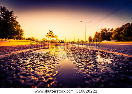 Sunset after rain, the parked car on the highway. Wide angle view close up with puddles on the asphalt level, image vignetting and the orange-purple toning