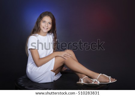 Pretty teen girl smiling while looking at the camera. Full length sitting in the studio on dark background with multicolored lights