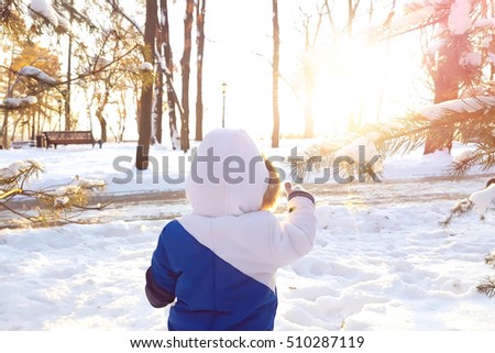 Year-old laughing baby boy in warm snowsuit walking in the winter park with a white snow. First winter and first toddler steps on the snow. Silhouette little child walking on a winter sunny day