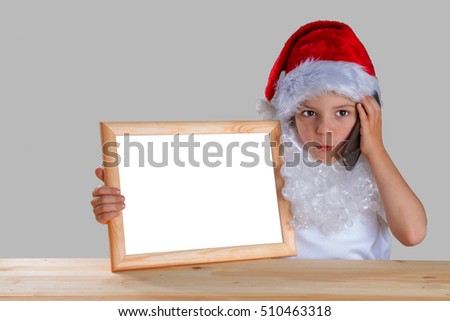Little Santa holding a blank picture frame with white background and talking on the phone. He is sitting at a wooden table looking at the camera. Close-up. Gray background.