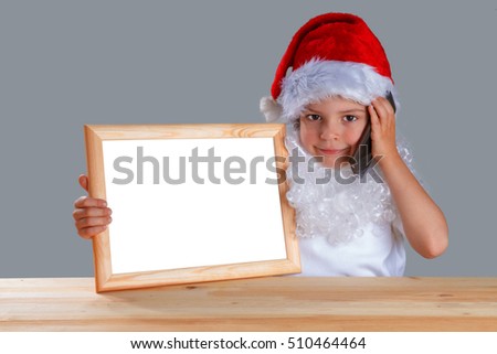 Little Santa holding a blank picture frame with a white background and a telephone. He is sitting at a wooden table looking at the camera, smiling. Close-up. Gray background.