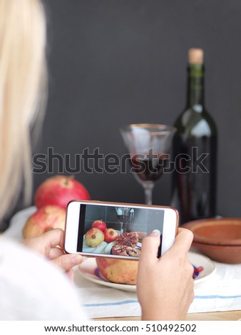 Photographer girl taking picture with a phone camera, of a still life with pomegranates and sweet wine bottle.