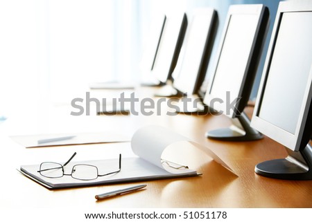 Close-up of workplace with notepad, pen, eyeglasses and monitors on it