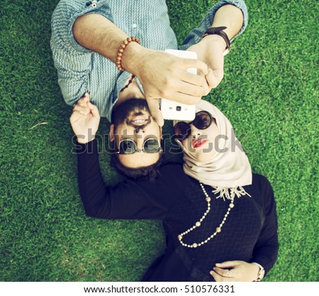 love and people concept - happy teenage arabic couple in sunglasses lying on grass and taking selfie on smartphone at summer