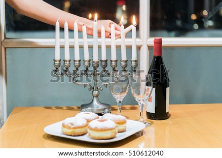 Low key image of Jewish holiday Hanukkah with menorah, sufganiyah jelly doughnuts and bottle of red wine with two glasses by the window with the night view on Tel Aviv, Israel.