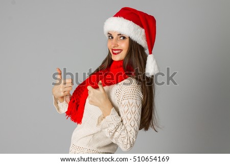 cheerful girl in santa hat and red scarf showing thumbs up in studio on gray background