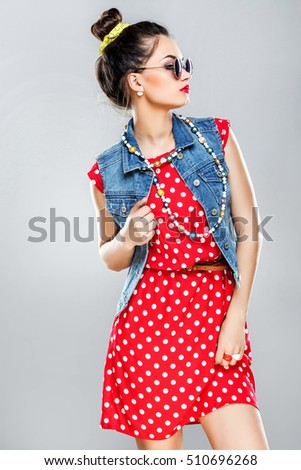Young girl with dark hair and red lips wearing red dress and sunglasses, posing at gray studio background, portrait.
