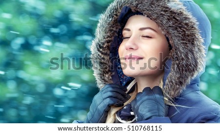 Portrait of a nice girl in winter park, with closed eyes enjoying falling snow, wearing warm coat with fur hood, happy wintertime holidays 