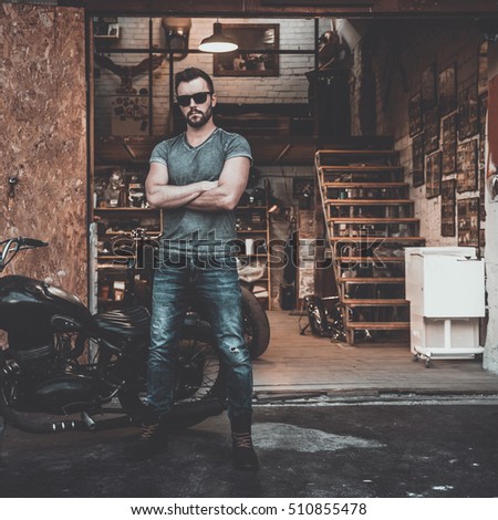 Welcome to my world! Full length of confident young man keeping arms crossed and looking at camera while standing near his motorcycle garage or repair shop