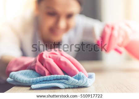 Woman in protective gloves is smiling and wiping dust using a spray and a duster while cleaning her house, close-up Royalty-Free Stock Photo #510917722