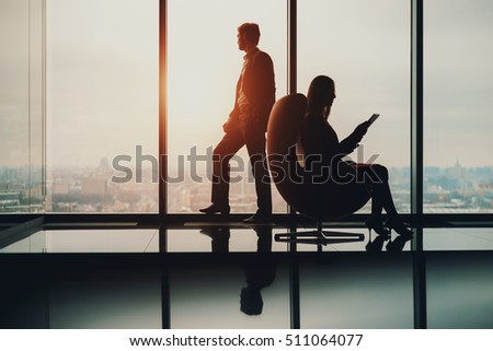 Silhouette of businessman standing next to big window, looking at blurred cityscape outside, his female colleague with digital tablet sitting on armchair and reading, office interior