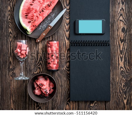 Smartphone on the blank black menu with watermelon slices, knife and glass.