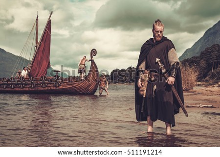 Viking warrior with sword standing near Drakkar on the seashore. Royalty-Free Stock Photo #511191214