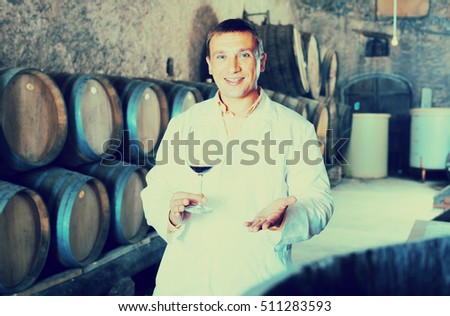 joyful factory sommelier in white robe checking quality of red wine in cellar with barrels
