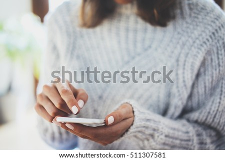 Closeup image of female hands using modern smartphone device at home interior, young hipster girl wearing cozy sweater and typing on touch screen of cellphone an sms message, social networking concept