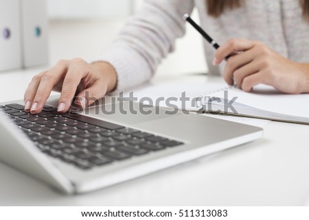 Close up of woman's hands. She is typing and holding a pen. White binders are seen at the background. Concept of office work. 