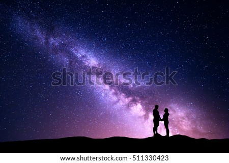 Milky Way with silhouette of people. Landscape with night sky with stars and standing man and woman holding hands on the mountain. Hugging couple against purple milky way. Beautiful galaxy. Universe.
