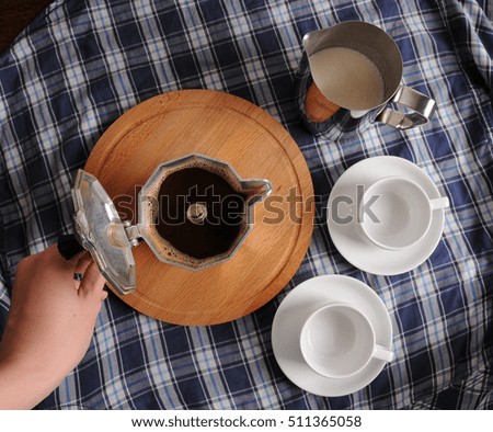 Geyser coffee on round wooden cutting board, a pitcher of milk and two white cups on blue plaid tablecloth