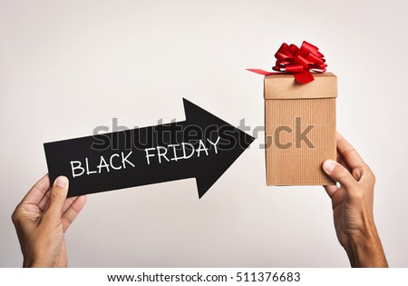 closeup of a young caucasian man holding a black arrow-shaped signboard with the text black friday written in it pointing to a gift box which holds in the other hand