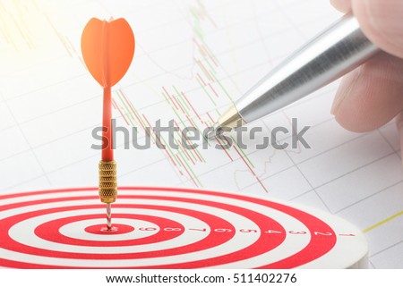 Red dart hits in the center of a target with a background of a hand analyses a technical chart of financial instruments. An idea of selecting the right asset for long term sustainable gain and growth.