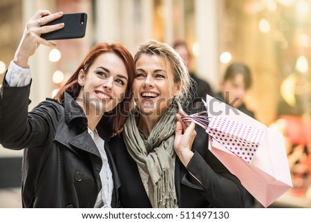 in the city, mother and daughter do shopping together, she takes the opportunity to make a selfie with a smartphone, lights of the shops at the background