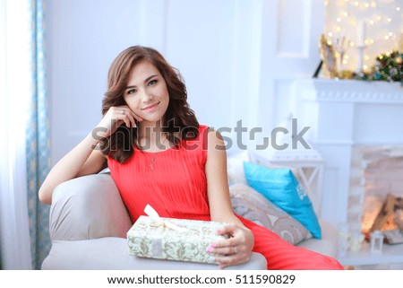 Young beautiful girl smiling, sitting on white couch and holding gift tied with beige ribbon in hands against window, white walls and lighted white stone fireplace. Girl dressed in long dress coral