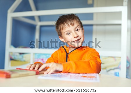 Boy is drawing with pencils. Smiling boy is  looking in the camera. Boy is in orange pullover. 