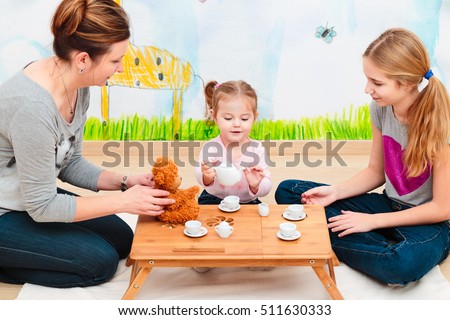 Little girl playing with her mother and elder sister at tea party using child's tea set