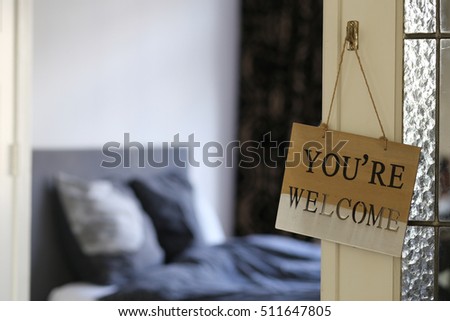 Hotel room, You are welcome words on wooden vintage frame card background, hanging on the bedroom door, enter the home. Welcoming hotel bed with pillows and backlight from the window. 