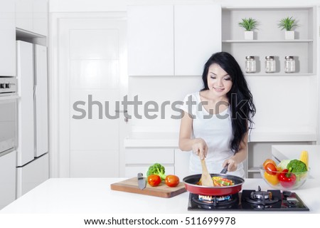Portrait of a beautiful Asian woman with long hair, cooking vegetable with a frying pan on the stove