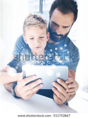 Bearded father with his young son using tablet PC in sunny room.Dad and little boy playing together on mobile computer, resting indoor.Childhood dreams icons concept.Vertical, blurred background
