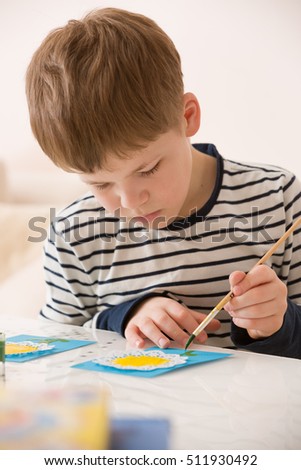 Portrait of little school boy sitting at the table and drawing with yellow paint. After school activities at home. Education. Small boy creating handmade cards.