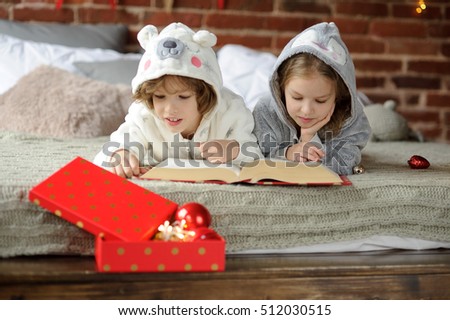 Christmas holiday. Two children in soft pajamas lie on a big bed and with enthusiasm read the huge book with Christmas fairy tales. The bright red box with Christmas-tree decorations is near.