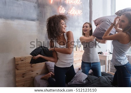 Laughing friends standing on the bed and having pillow fight