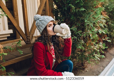 Portrait pretty girl with long hair in red coat, knitted hat and white gloves sitting on wooden stairs outdoor. She drinks coffee and looking to camera
