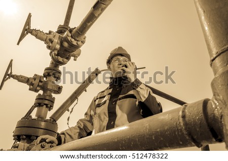 Oilfield worker near wellhead valve, wearing helmet and work clothes talking on the radio. Oil and gas concept. Toned.