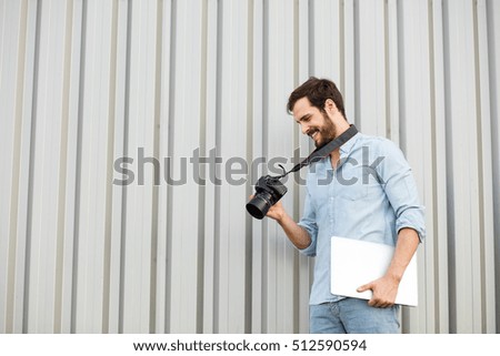 photographer looking at his camera with laptop in hands on gray wall