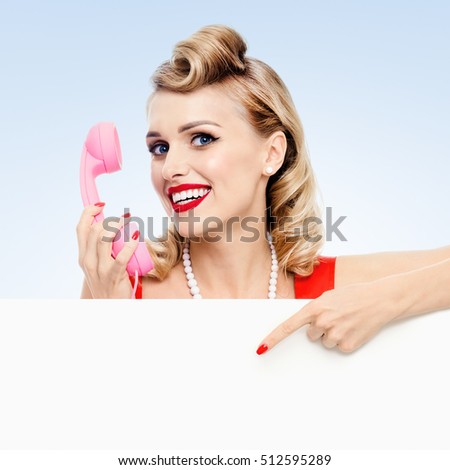 Woman with phone, in pin-up style dress, showing blank signboard with copyspace area, on blue background. Caucasian blond model posing in retro fashion and vintage concept studio shoot.