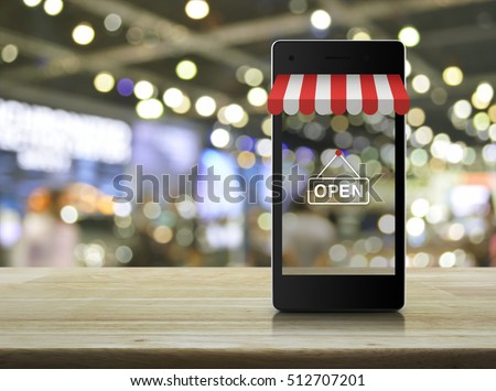 Modern smart mobile phone with on line shopping store graphic and open sign on wooden table over blur light and shadow of mall
