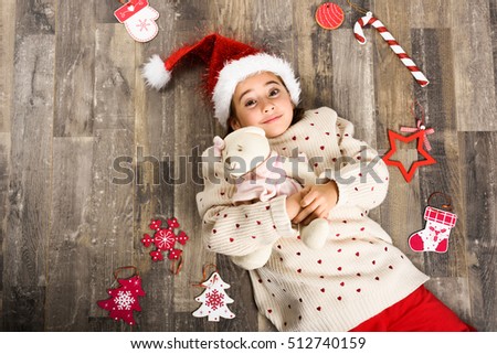 Adorable little girl wearing santa hat smiling on wooden floor with Christmas ornaments. Winter clothes.