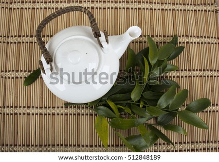 White teapot with green leaves on bamboo mat plate. Top view
