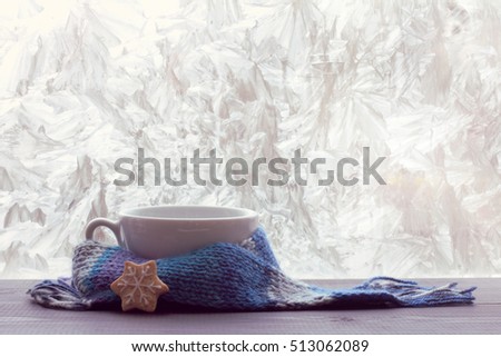 White empty mug for warming beverage wrapped in a scarf on a table next to a frosty winter window / time pour hotter