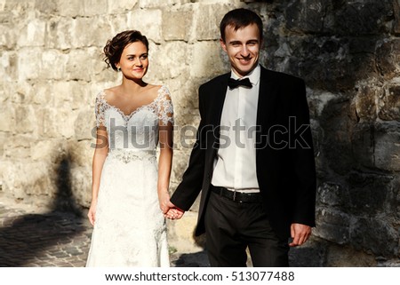 Happy groom holds bride's hand standing in the rays of morning sun